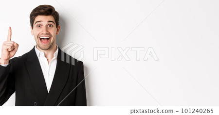 Close-up of handsome businessman in black suit, smiling amazed, showing number one, standing over white background 101240265