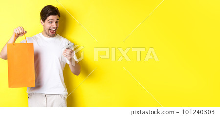 Concept of discounts, online banking and cashback. Surprised man showing shopping bag and looking happy at mobile screen, standing against yellow background 101240303