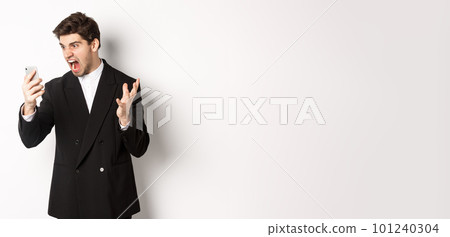 Portrait of angry businessman in black suit yelling at mobile phone, having an argument on video call, standing mad over white background 101240304