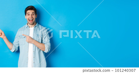 Excited handsome 25s years man with beard, pointing fingers left and smiling amazed, standing against blue background 101240307