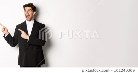 Portrait of amazed and happy handsome man in black suit, pointing and looking left impressed, showing awesome christmas promo, standing over white background Portrait of amazed and happy handsome man in black suit, pointing and looking left impressed, showing awesome christmas promo, standing over white background 101240309