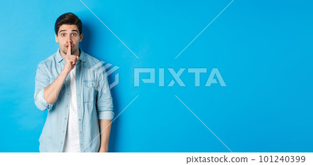 Portrait of excited man asking to keep quiet, showing hush taboo sign and looking nervously at camera, standing against blue background 101240399