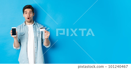 Amazed young man showing mobile cell phone screen and credit card, shop online, standing against blue background Amazed young man showing mobile cell phone screen and credit card, shop online, standing against blue background 101240467