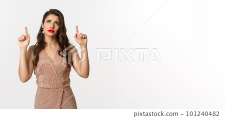 Christmas, holidays and celebration concept. Elegant woman in luxury dress looking doubtful at logo, pointing fingers up, standing over white background 101240482