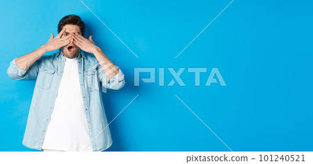 Shocked man covering eyes and peeking through fingers, stare at something embarrassing, standing against blue background 101240521