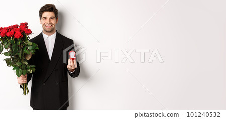 Handsome smiling man in black suit, holding roses and engagement ring, making a proposal to marry him, standing against white background 101240532