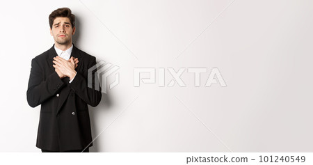 Portrait of touched and compassionate guy in suit, holding hands on heart and looking with pity at camera, standing over white background Portrait of touched and compassionate guy in suit, holding hands on heart and looking with pity at camera, standing over white background 101240549