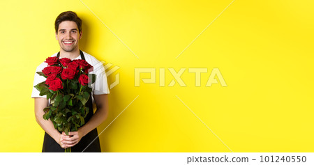 Smiling florist in black apron holding flowers, selling bouquet of roses, standing over yellow background Smiling florist in black apron holding flowers, selling bouquet of roses, standing over yellow background 101240550