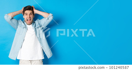 Frustrated guy losing, yelling displeased and looking at camera, holding hands on head, standing over blue background 101240587