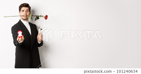 Romantic young man in suit making a proposal, holding rose in teeth and glass of champagne, showing engagement ring, asking to marry him, standing against white background Romantic young man in suit making a proposal, holding rose in teeth and glass of champagne, showing engagement ring, asking to marry him, standing against white background 101240634