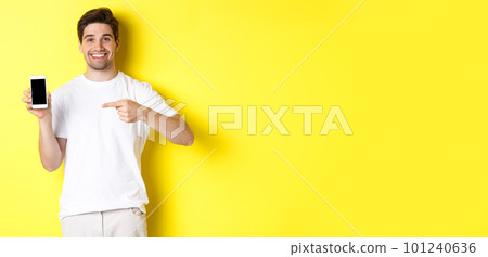 Image of attractive young man pointing finger at smartphone screen, showing an app, standing against yellow background Image of attractive young man pointing finger at smartphone screen, showing an app, standing against yellow background 101240636