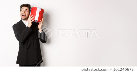 Concept of christmas holidays, celebration and lifestyle. Image of excited man enjoying new year, shaking gift box to guess what inside, standing against white background Concept of christmas holidays, celebration and lifestyle. Image of excited man enjoying new year, shaking gift box to guess what inside, standing against white background 101240672