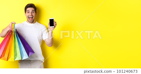 Young man holding shopping bags and showing mobile phone screen, money application, standing over yellow background Young man holding shopping bags and showing mobile phone screen, money application, standing over yellow background 101240673