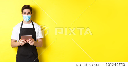 Concept of covid-19, small business and pandemic. Waiter in black apron and medical mask taking order, holding digital tablet, standing over yellow background Concept of covid-19, small business and pandemic. Waiter in black apron and medical mask taking order, holding digital tablet, standing over yellow background 101240750
