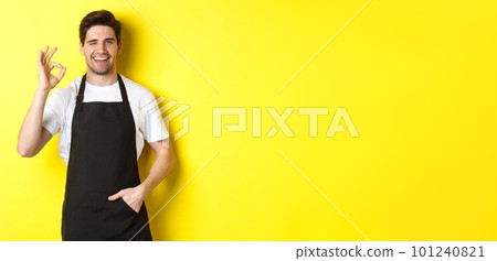 Confident and handsome waiter showing ok sign, wearing black apron and standing against yellow background 101240821