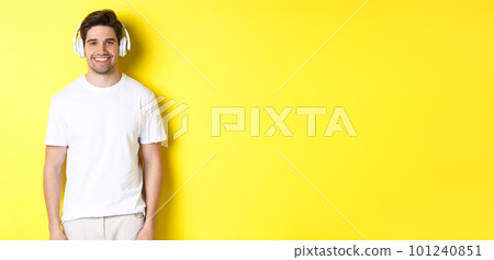 Young handsome man listening music in headphones, wearing earphones and smiling, standing over yellow background 101240851