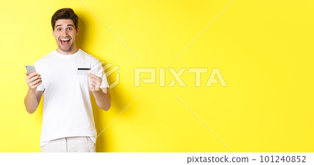 Happy male buyer holding smartphone and credit card, concept of online shopping in internet, standing over yellow background 101240852