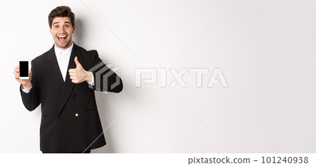Image of handsome male entrepreneur in black suit, recommending app or online shop, showing thumbs-up and smartphone screen, standing over white background 101240938