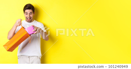 Concept of holidays and celebration. Young man looking surprised as take out gift from shopping bag, standing over yellow background 101240949