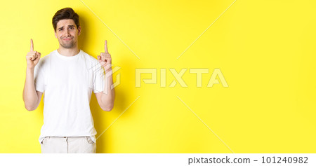 Unamused handsome guy frowning, pointing fingers up at something bad, standing skeptical against yellow background Unamused handsome guy frowning, pointing fingers up at something bad, standing skeptical against yellow background 101240982