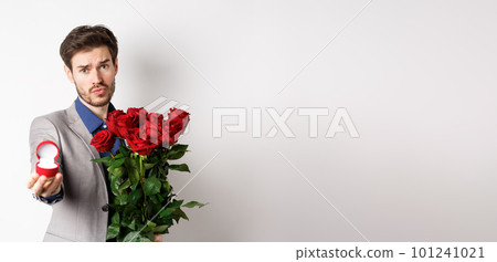 Handsome boyfriend in suit asking to marry him, standing with red bouquet of roses and engagement ring, looking pleading at camera, standing over white background Handsome boyfriend in suit asking to marry him, standing with red bouquet of roses and engagement ring, looking pleading at camera, standing over white background 101241021