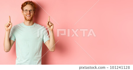Attractive young man in glasses and t-shirt showing promo deal, pointing fingers up and smiling at camera, standing over pink background Attractive young man in glasses and t-shirt showing promo deal, pointing fingers up and smiling at camera, standing over pink background 101241128