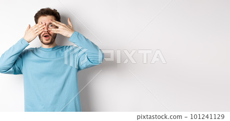 Amazed smiling man covering eyes with hands but peeking through fingers at something awesome, standing over white background Amazed smiling man covering eyes with hands but peeking through fingers at something awesome, standing over white background 101241129