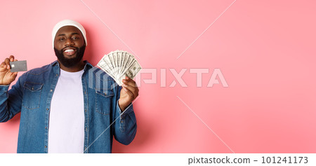 Shopping concept. Handsome young Black guy showing credit card of bank and money, smiling satisfied, standing over pink background Shopping concept. Handsome young Black guy showing credit card of bank and money, smiling satisfied, standing over pink background 101241173