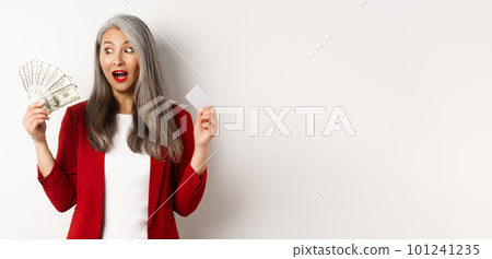 Surprised asian middle-aged woman with grey hair, looking at money and holding credit card, earning cash, standing over white background Surprised asian middle-aged woman with grey hair, looking at money and holding credit card, earning cash, standing over white background 101241235