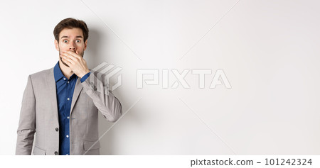 Shocked boyfriend in suit gasping, covering mouth with hand and pop eyes at camera startled, standing on white background 101242324