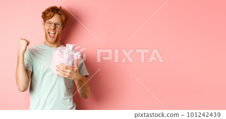 Valentines day and romance concept. Cheerful redhead guy making fist pump and scream yes with joy, holding gift box, smiling, standing over pink background Valentines day and romance concept. Cheerful redhead guy making fist pump and scream yes with joy, holding gift box, smiling, standing over pink background 101242439