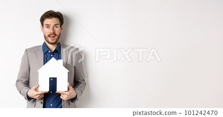 Real estate. Excited business man in suit holding paper house cutout and looking amazed at camera, buying apartment, white background Real estate. Excited business man in suit holding paper house cutout and looking amazed at camera, buying apartment, white background 101242470