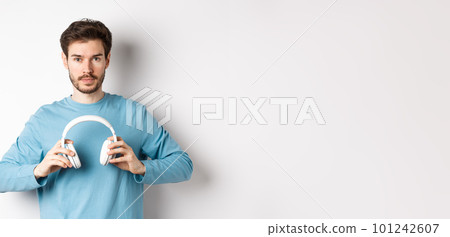 Young bearded guy in blue sweatshirt put on wireless headphones, listening music, standing on white background Young bearded guy in blue sweatshirt put on wireless headphones, listening music, standing on white background 101242607