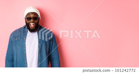 Surprised and happy african-american hipster man in beanie and sunglasses, smiling and looking at camera, standing confident over pink background Surprised and happy african-american hipster man in beanie and sunglasses, smiling and looking at camera, standing confident over pink background 101242772