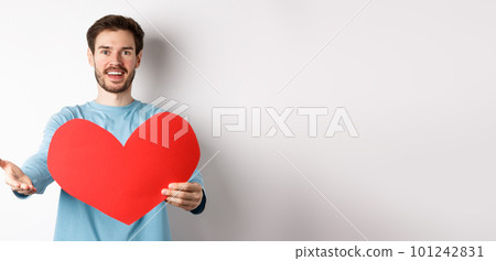 Handsome man in love making confession to you, pointing hand at camera, holding big red heart cutout on valentines day, singing romantic serenade, standing over white background 101242831
