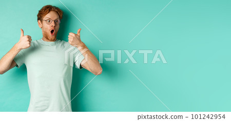 Excited young man with red hair and beard, staring at promo with awe, showing thumbs-up in approval, praising offer, standing over turquoise background Excited young man with red hair and beard, staring at promo with awe, showing thumbs-up in approval, praising offer, standing over turquoise background 101242954