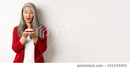 Image of funny asian senior woman looking tempted at piece of cake, desire to bite dessert, standing over white background 101243003