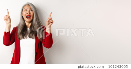 Happy asian lady in red blazer and makeup, looking and pointing fingers up, showing special offer, standing over white background 101243005