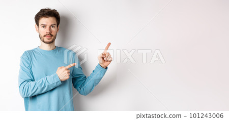 Portrait of serious caucasian man with beard pointing right at logo and looking at camera, standing in casual clothes on white background 101243006