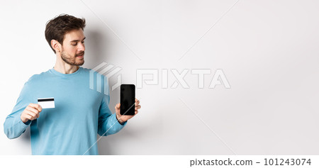 Mobile banking. Handsome caucasian man showing plastic credit card and looking at blank mobile screen, standing over white background Mobile banking. Handsome caucasian man showing plastic credit card and looking at blank mobile screen, standing over white background 101243074