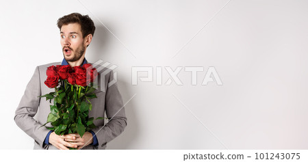Handsome young man in suit holding red roses, looking left with surprised and startled expression, standing on Valentines day over white background 101243075