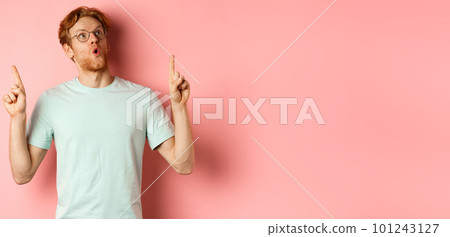 Wow check this out. Surprised redhead guy in glasses looking and pointing up, gasping amazed, standing over pink background 101243127