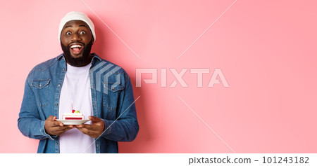 Cheerful african-american guy celebrating birthday, making wish on bday cake with lit candle, smiling happy, standing over pink background 101243182