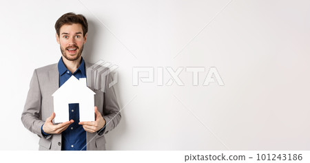 Real estate. Excited male entrepreneur holding paper house cutout, wearing suit and look hopeful at camera 101243186