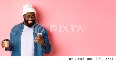 Image of stylish Black man hipster drinking takeaway coffee, reading message on phone and smiling, standing over pink background 101243351