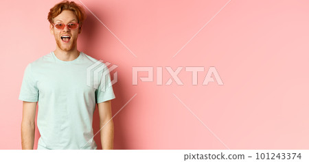 Image of surprised redhead man on vacation, wearing sunglasses with summer t-shirt, open mouth and saying wow amazed, standing over pink background 101243374