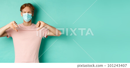 Covid-19, pandemic and lifestyle concept. Young man with red hair in face mask, showing thumbs down, dislike or disapprove something, standing over turquoise background 101243407