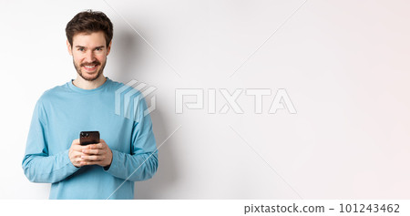 Smiling modern man using smartphone and looking pleased at camera. Guy in sweatshirt with mobile phone on white background 101243462