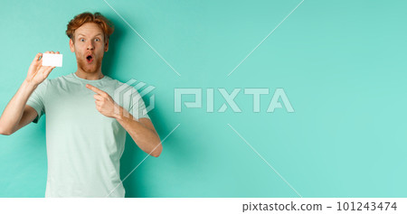 Shopping concept. Handsome redhead man in t-shirt showing plastic credit card and smiling, standing over turquoise background 101243474