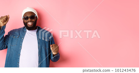 Happy Black man in beanie an sunglasses rejoicing, dancing with happy face, standing over pink background 101243476